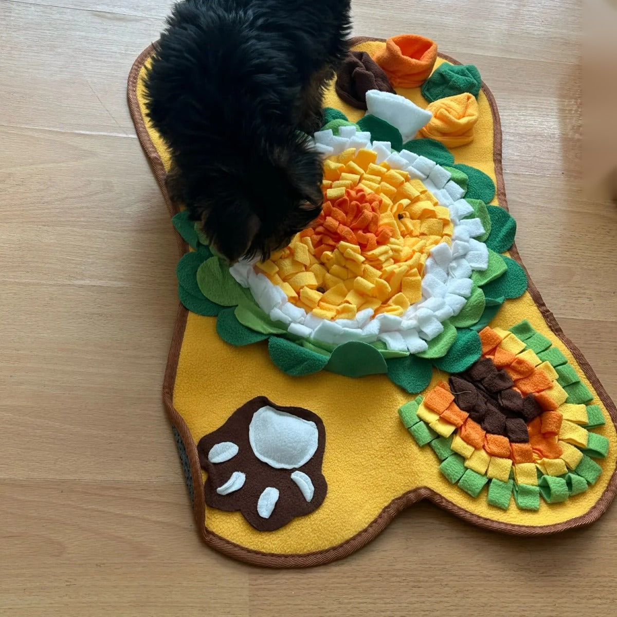Yellow bone-shaped fleece snuffle mat (64 x 43 cm) with a big layered flower, paw-print patch and veg patch, with a small dog sniffing for treats